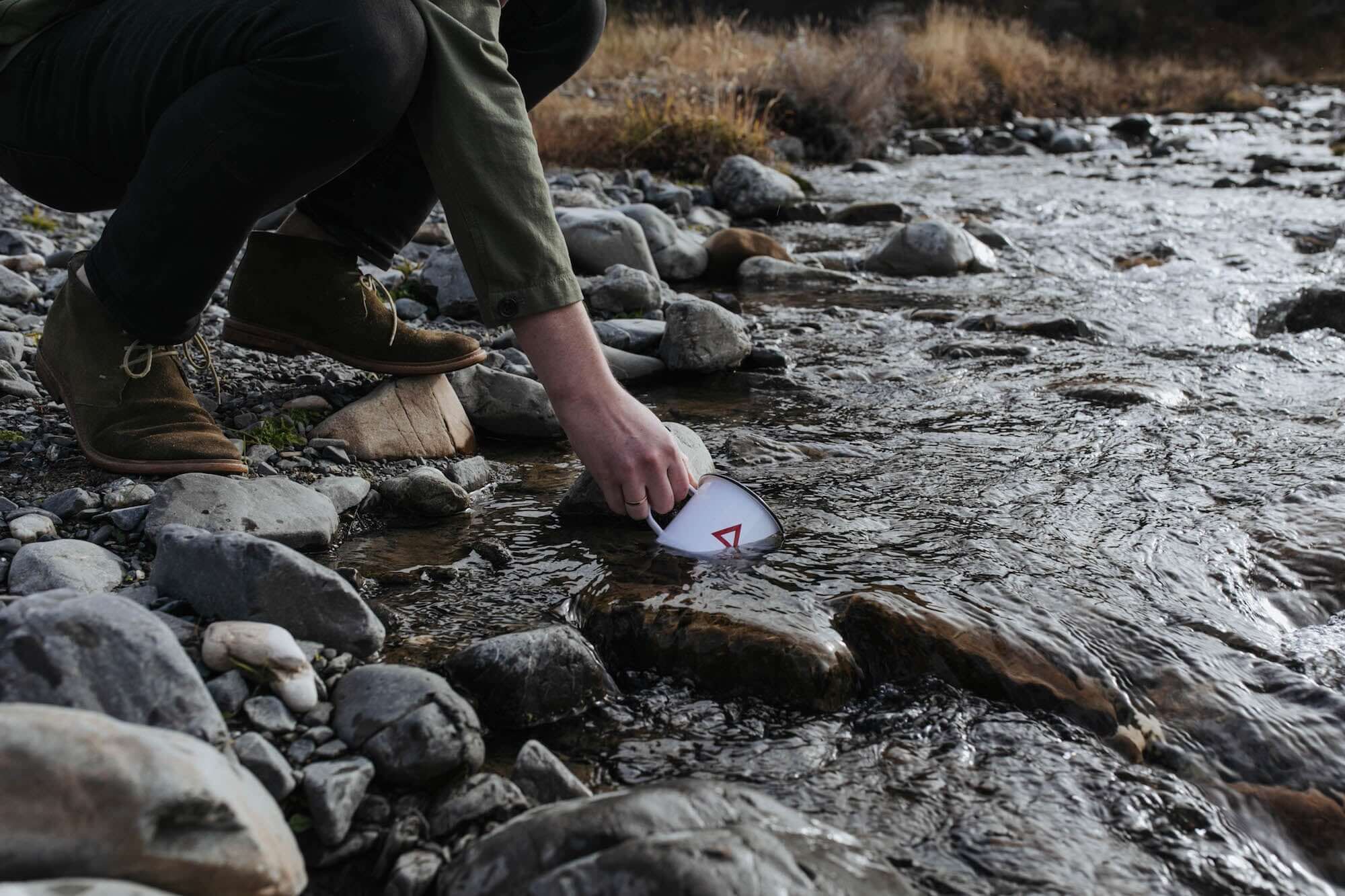 Person picking up a Classic Enamel Mug from a river, surrounded by rocks and flowing water.