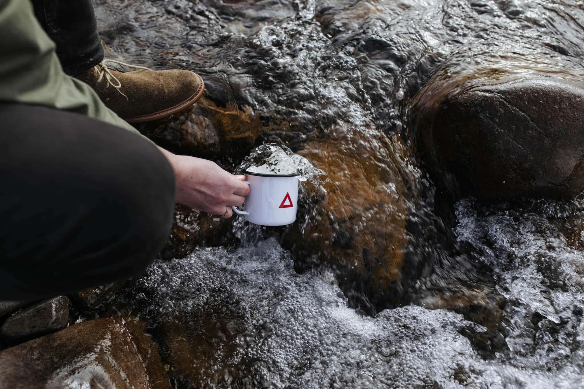 Person holding a Classic Enamel Mug near a flowing stream, showcasing its durable design and outdoor appeal.